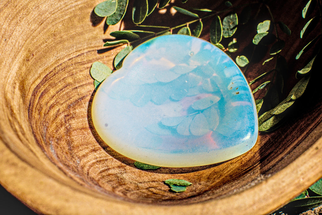 A smooth, polished opalite stone with a mskyblue softness, white, and soft pink orange light filtering, resting on a wooden bowl with green leaves.